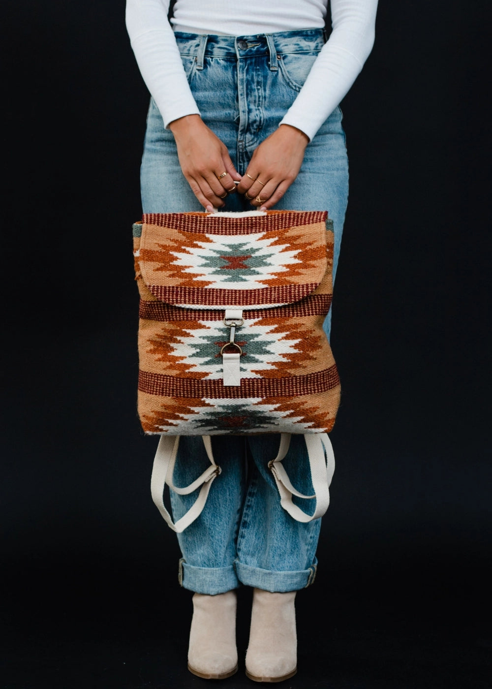 A person wearing cuffed blue jeans and beige suede boots holds a woven backpack with a Southwestern-style geometric pattern in rust orange white and teal against a black background