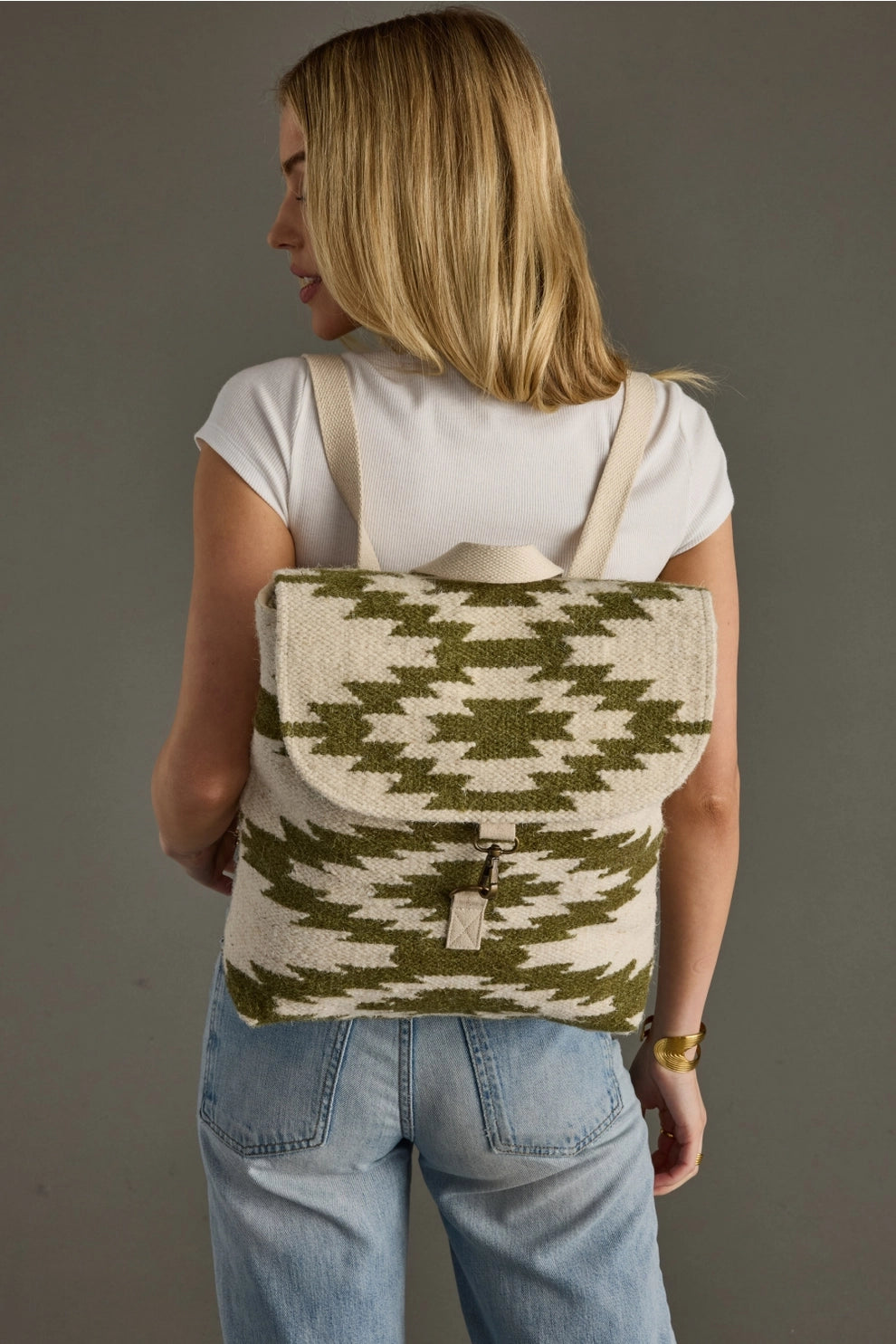 A blonde woman from behind models a cream and olive green woven geometric pattern backpack with canvas straps, wearing a white ribbed t-shirt, light wash jeans, and a gold cuff bracelet against a grey studio background