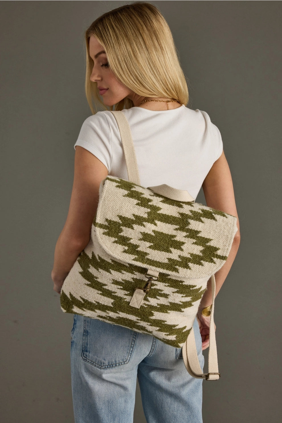A blonde woman models a cream and olive green geometric woven backpack with a distinctive staggered pattern and metallic clasp against a plain gray studio wall