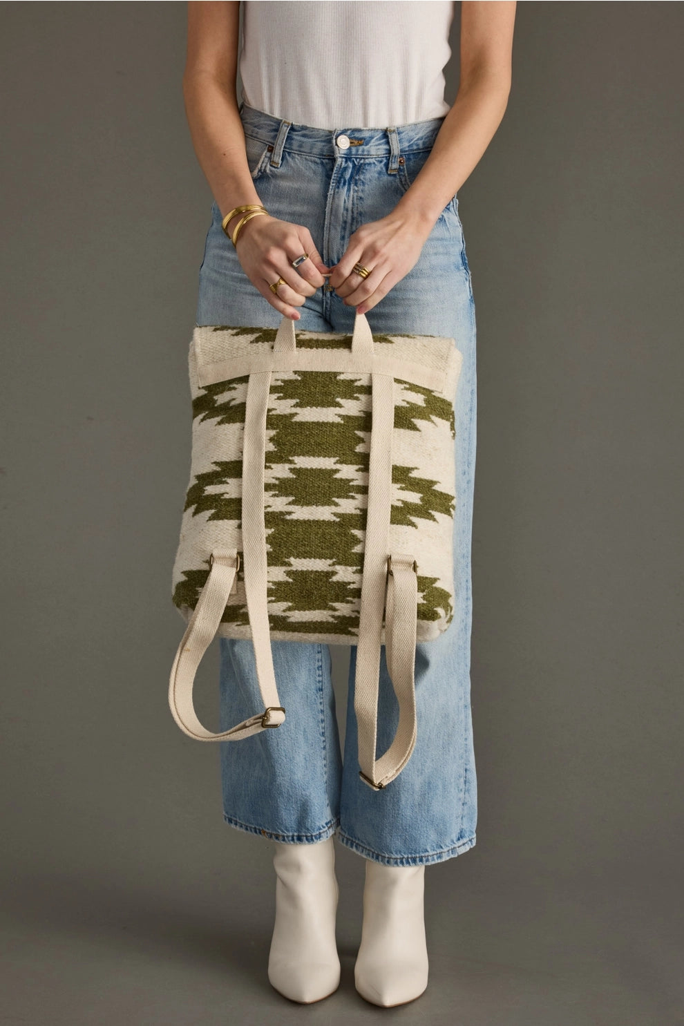 A woman wearing a white ribbed tank top, light wash wide-leg jeans, and white pointy-toe ankle boots holds a distinctive cream and olive green woven backpack featuring geometric patterns and beige canvas straps in a neutral studio