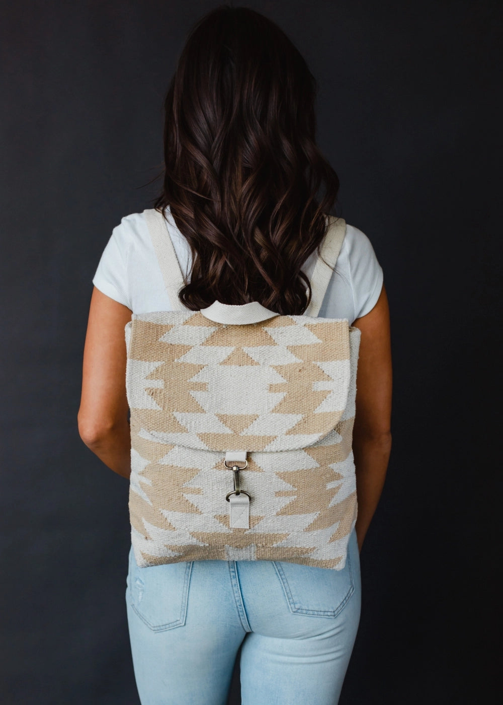 Photography of a woman's back in a studio setting wearing a cream and beige geometric patterned woven backpack with a silver metal clasp, showcasing her long dark wavy hair and light blue jeans