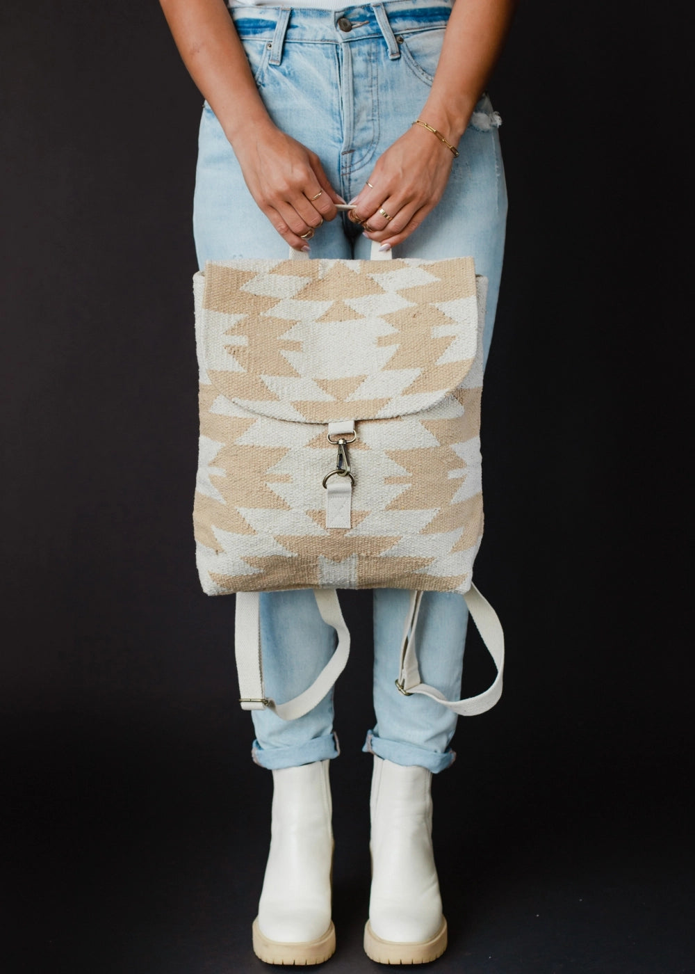 A person wearing light wash denim jeans, white chunky sole ankle boots, and gold jewelry holds a beige and off-white geometric patterned backpack with a silver hook clasp against a black studio backdrop