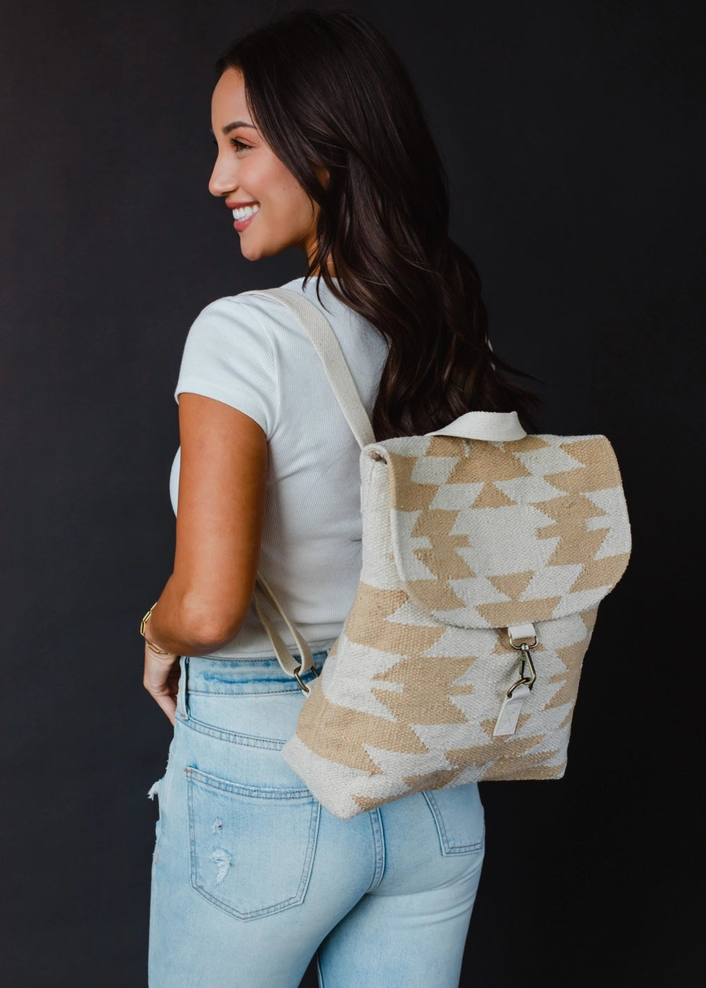A smiling woman with long dark hair wearing a white ribbed t-shirt, light wash distressed jeans, and a beige and cream geometric patterned backpack against a black background