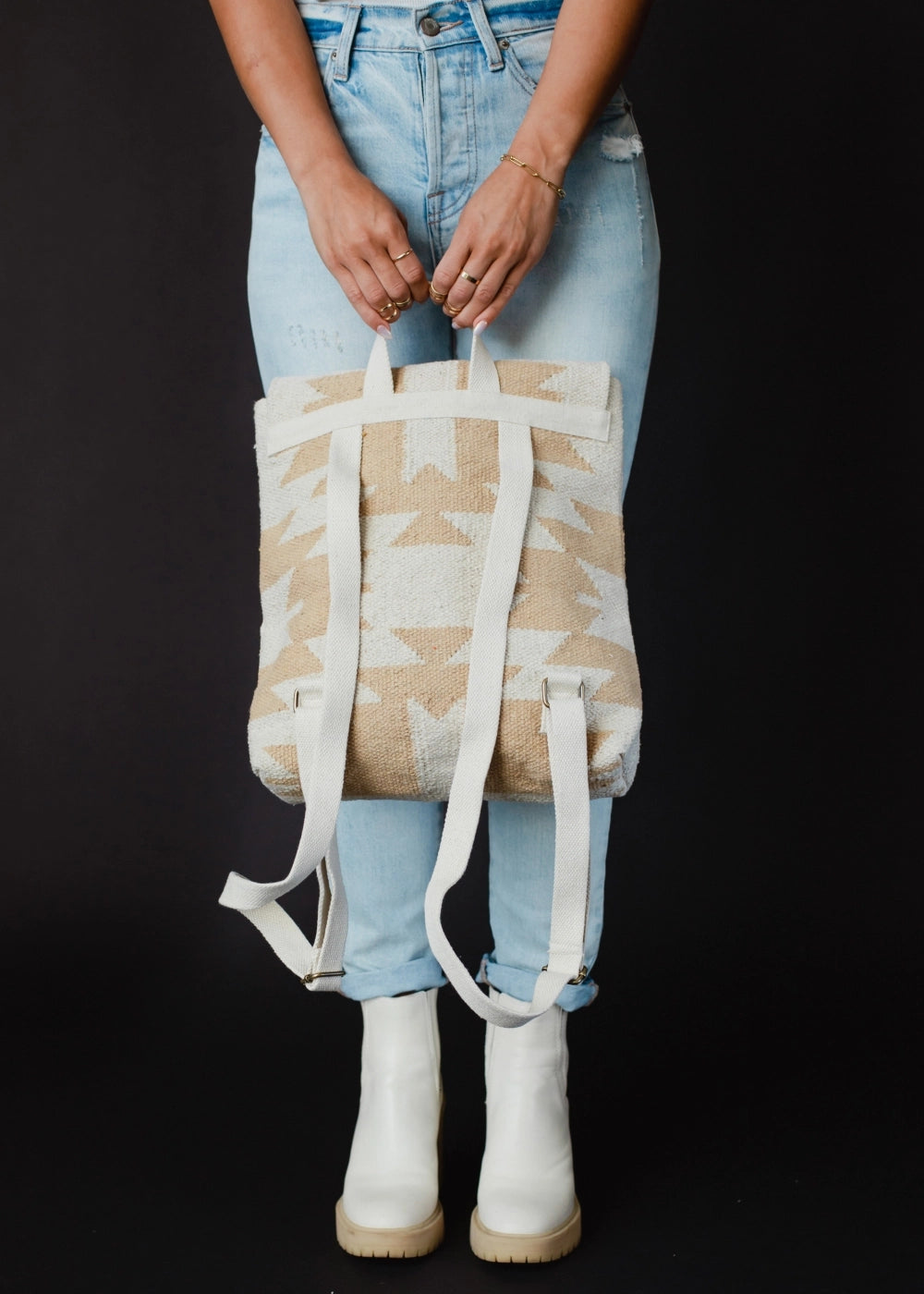 A person wearing light wash ripped jeans and white platform ankle boots with gold rings holds a tan and off-white geometric pattern backpack on a black background