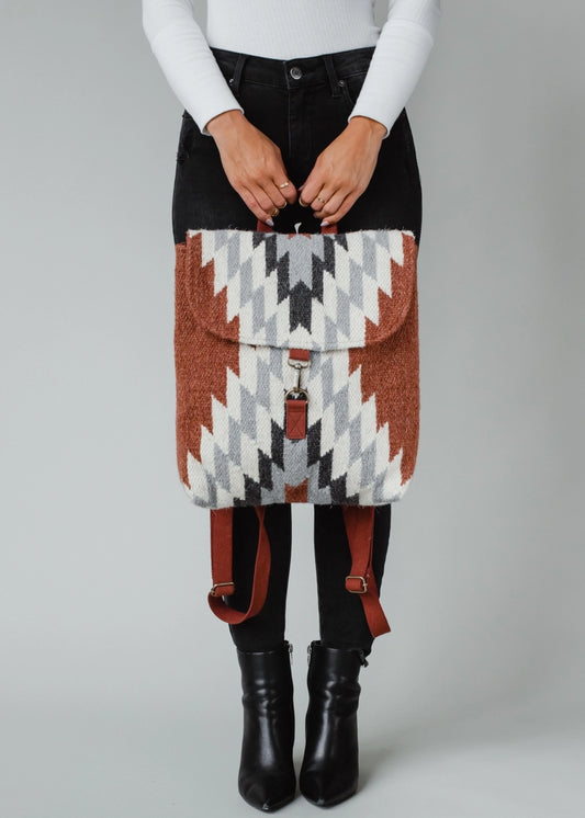 A person wearing black jeans and a white shirt holds a handcrafted backpack featuring a striking geometric pattern in earthy rust, white, grey, and black with visible brown leather straps and metal clasp