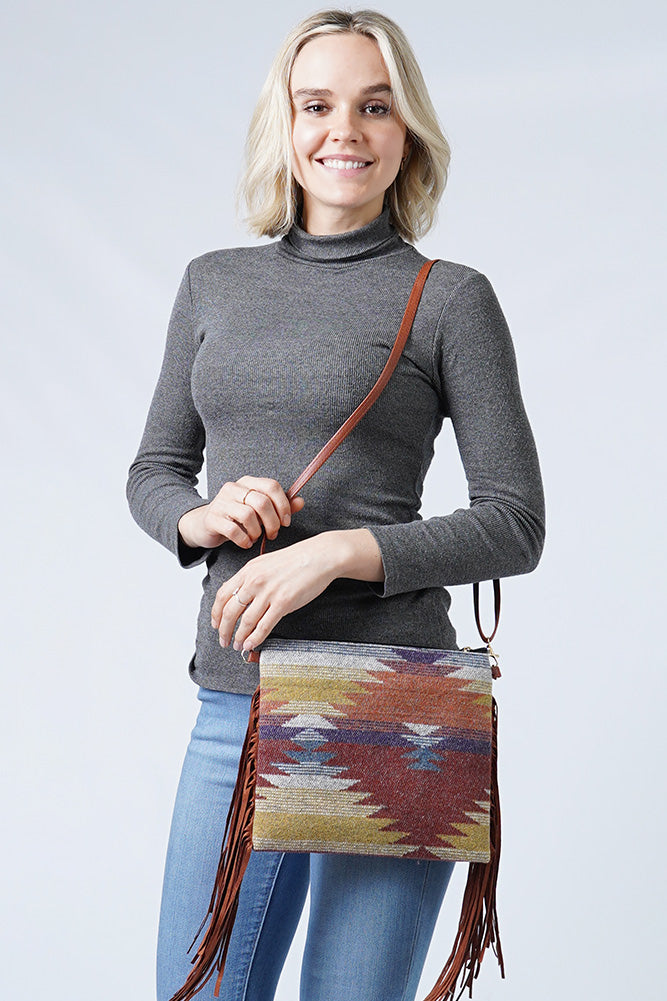 A smiling blonde woman in a grey turtleneck and blue jeans poses with a vibrant crossbody bag featuring a Southwestern geometric pattern and brown leather fringe
