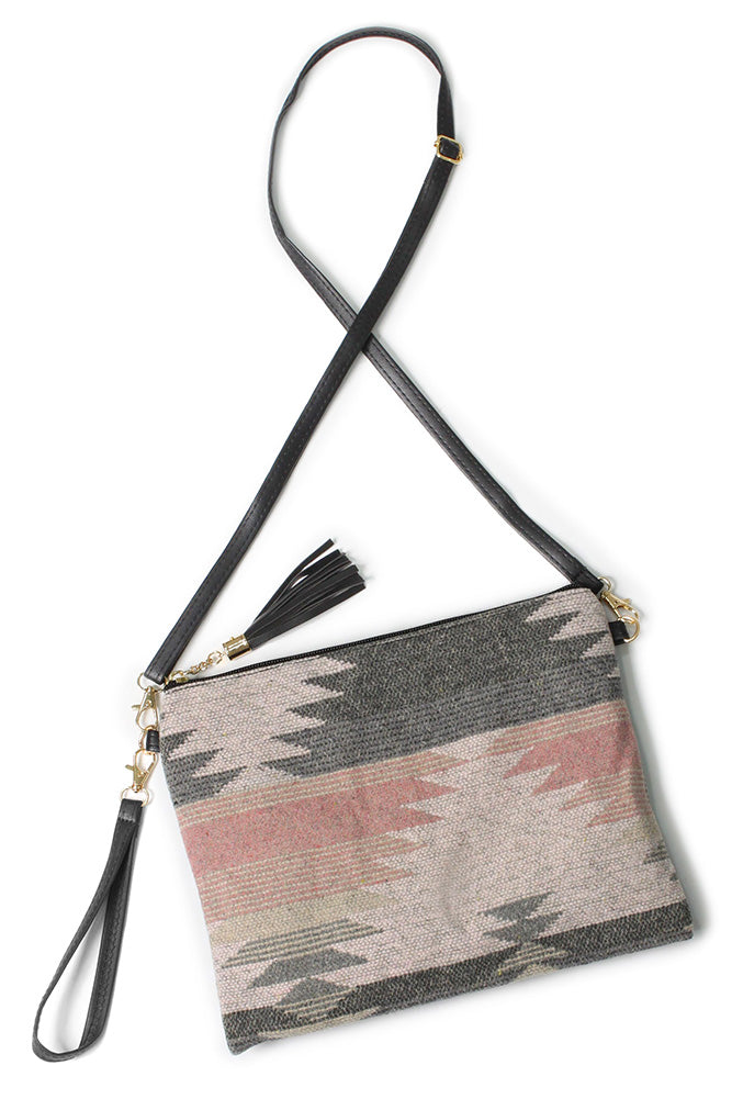 Woven fabric crossbody clutch bag featuring a geometric Southwestern pattern in shades of gray dusty pink and beige with black leather straps and tassel detail accented by gold hardware
