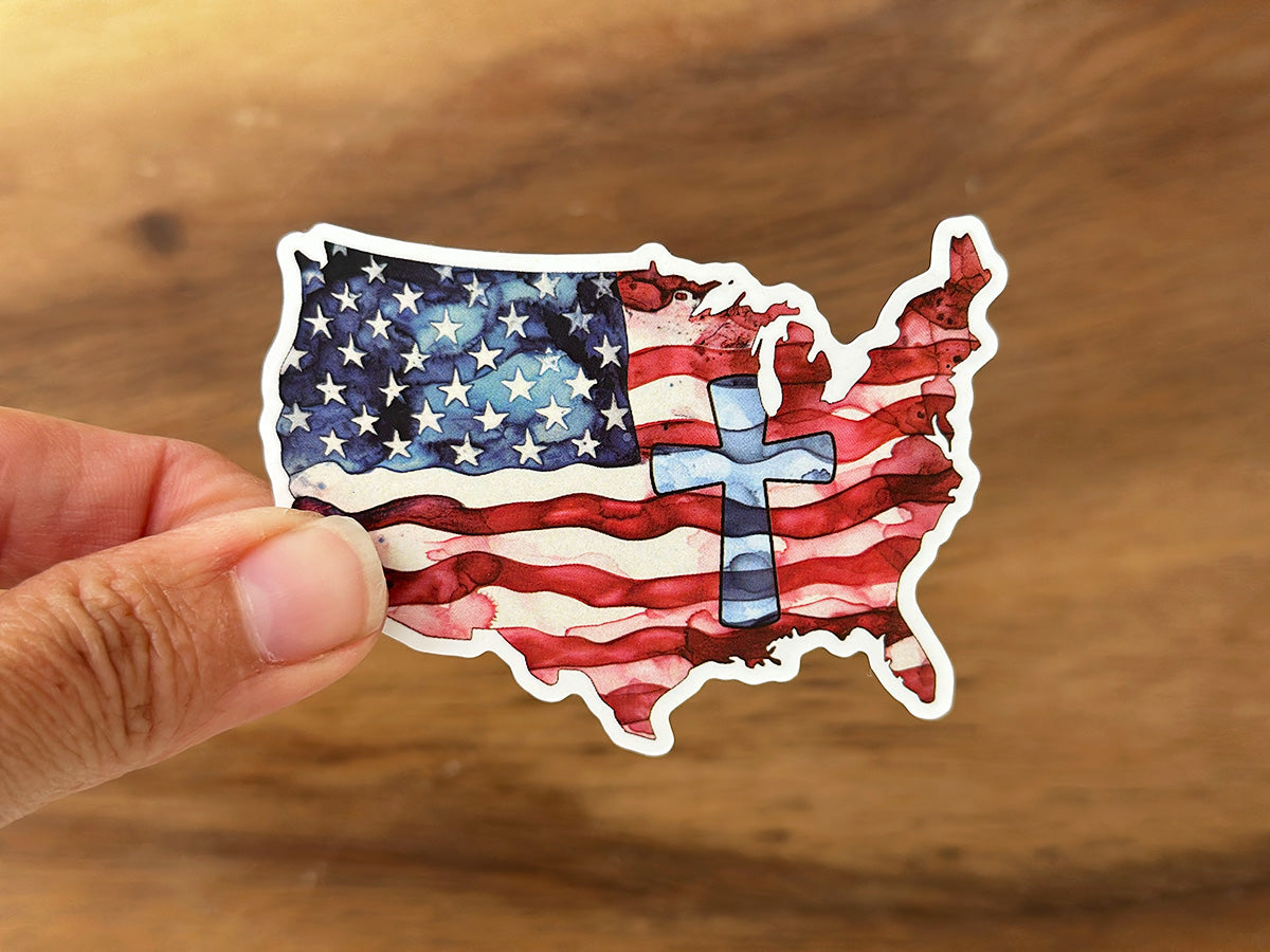 Watercolor art sticker depicting the contiguous United States map as an American flag with vibrant red stripes, a blue star field, and a transparent blue cross centrally overlaid, held by a hand against a wood background