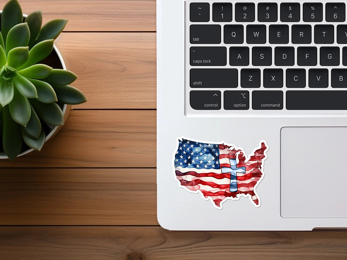 Photograph of a flat lay desktop setup featuring a silver laptop with a United States map sticker showing a watercolor American flag design and a prominent blue cross, next to a vibrant green succulent plant on a warm wooden desk surface