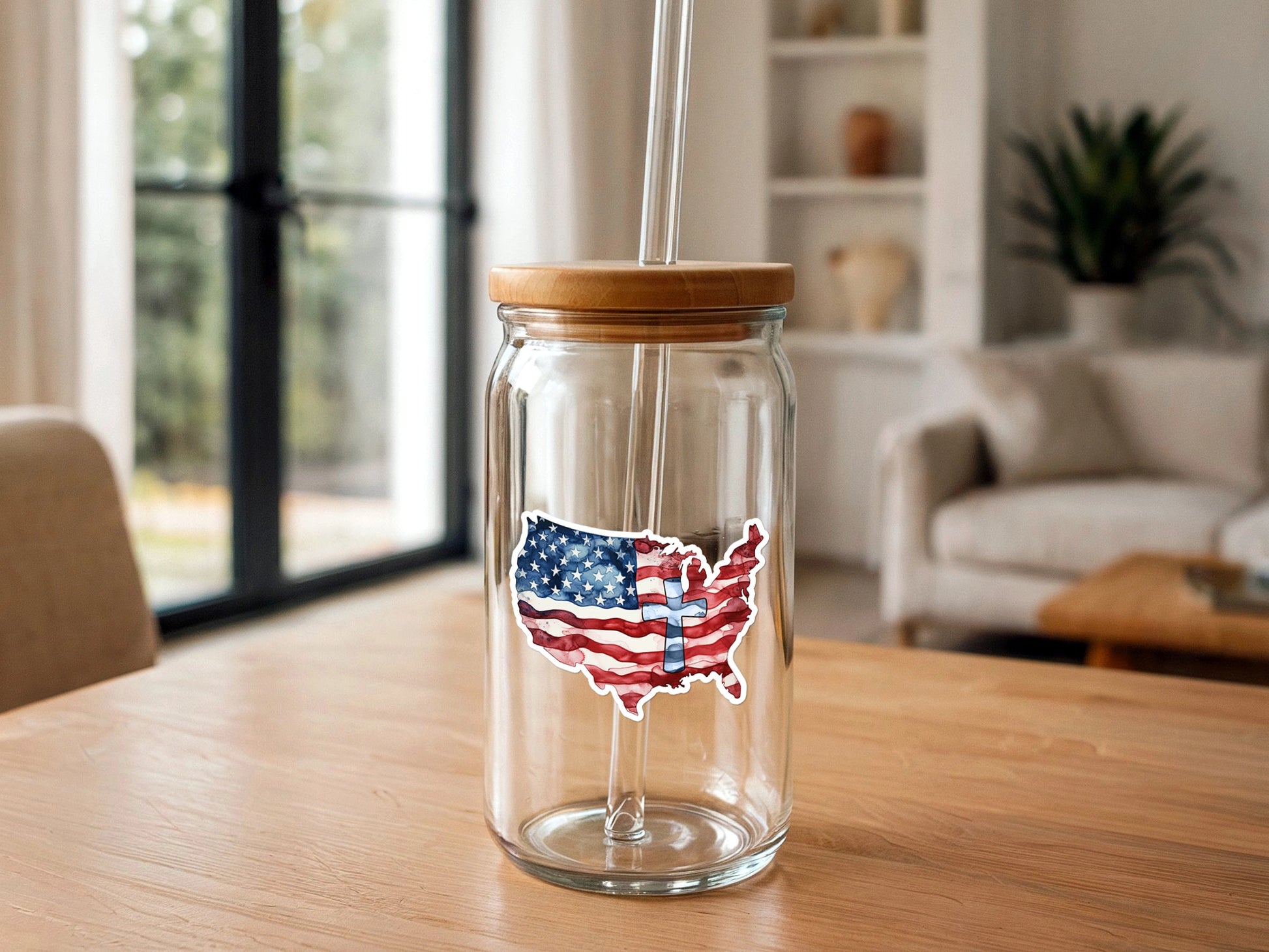 Photograph of a clear glass tumbler with a wooden lid and glass straw, adorned with a watercolor US map sticker in American flag colors featuring a light blue cross, on a light wood table in a modern sunlit room