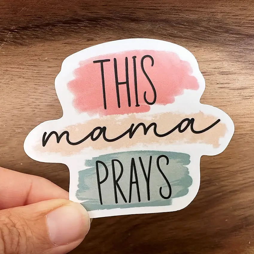 Die-cut sticker held in a hand featuring the phrase THIS mama PRAYS in black text with watercolor washes of soft pink, pale beige, and light teal on a warm wooden background
