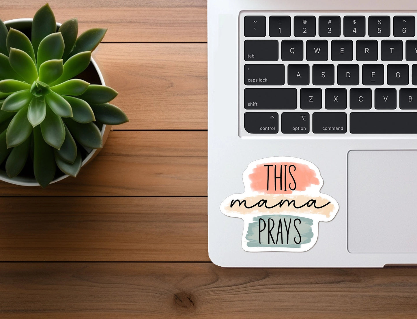 Overhead view of a modern desktop workspace featuring a vibrant green succulent plant in a white pot, a white laptop with black keys, and a colorful "THIS mama PRAYS" sticker with peach, tan, and teal watercolor swatches on a rich wooden desk surface