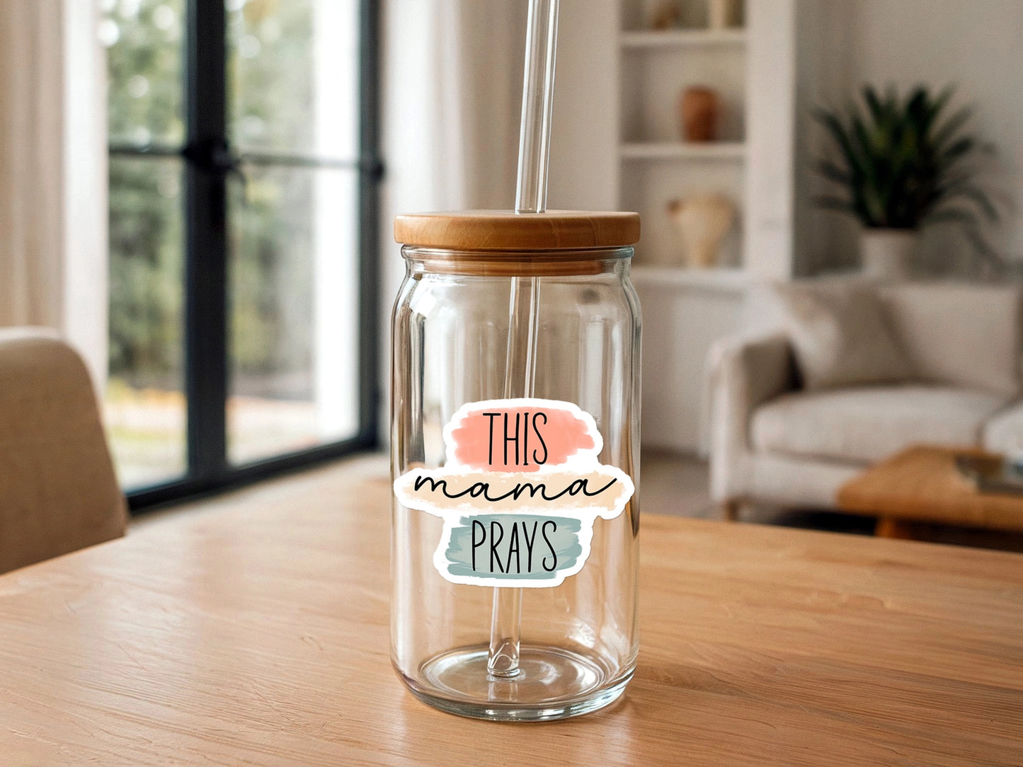 A clear glass can-shaped cup with a bamboo lid and glass straw displays a "THIS mama PRAYS" sticker with peach, tan, and teal watercolor backgrounds, positioned on a light wooden table against a blurry modern living room background featuring a large window and white couch