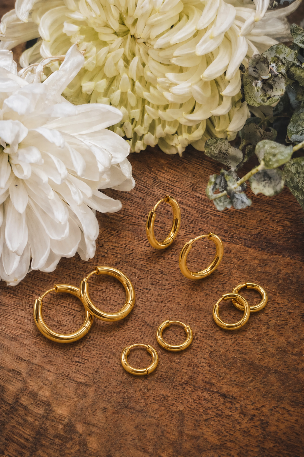 Photograph of six polished gold hoop earrings in assorted sizes scattered on a warm brown wooden surface, adorned with white and light green chrysanthemums and dark green eucalyptus foliage
