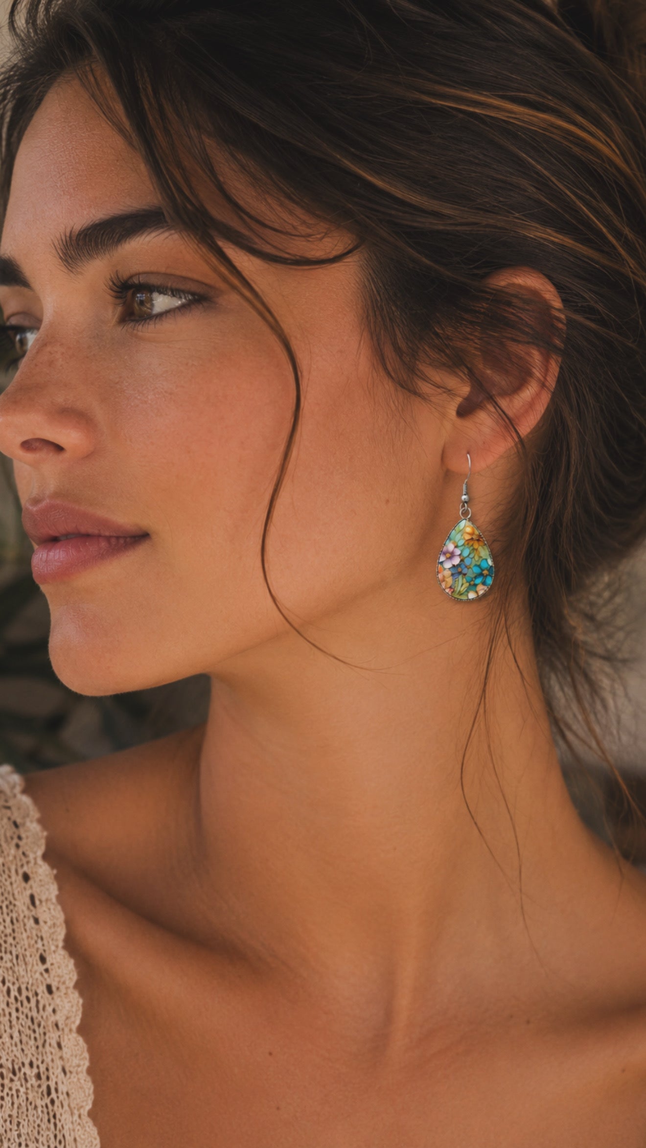 Photograph of a woman in profile with warm skin and dark hair wearing a vibrant floral teardrop earring and a delicate lace top against a softly blurred natural background