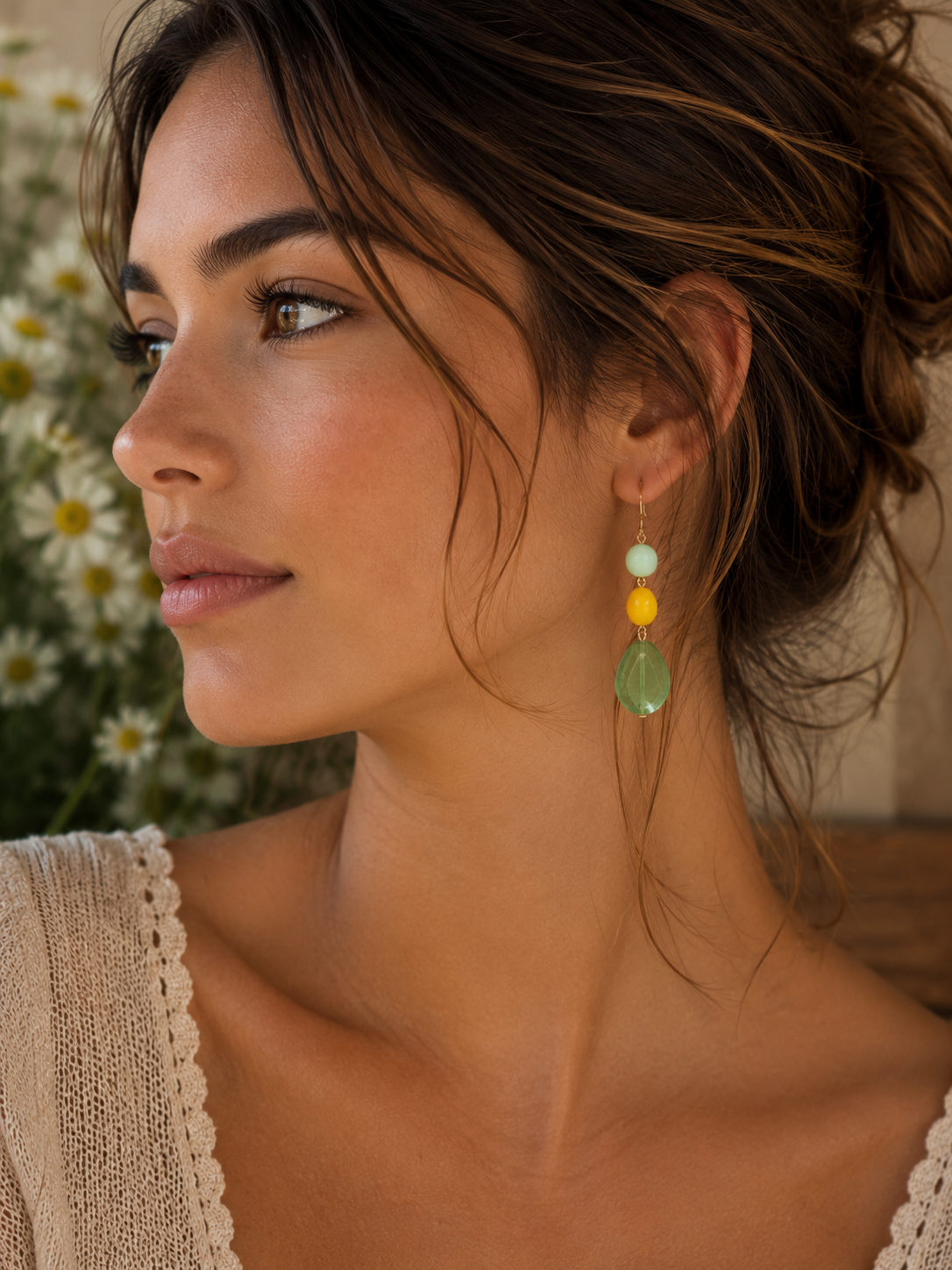 Photography of a woman in profile looking left wearing a pale green yellow and light blue beaded earring with a large green teardrop against a blurred background of white daisy flowers and a beige crochet knit top