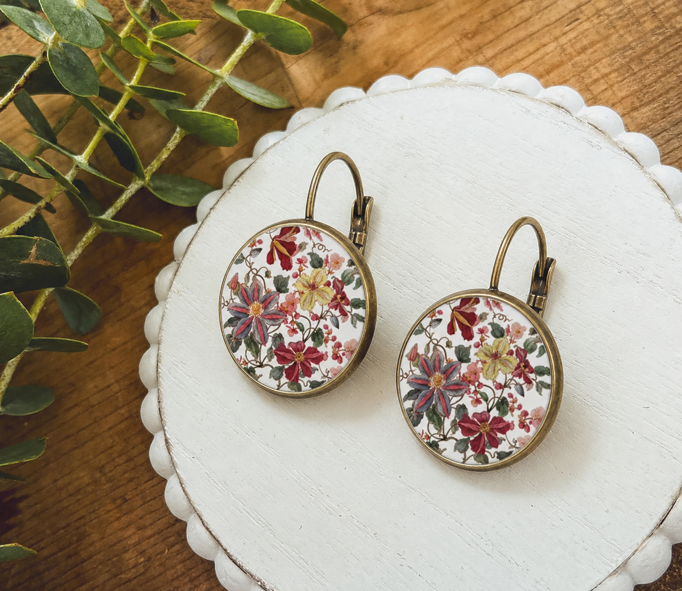 Pair of round antique bronze lever-back earrings featuring an intricate red yellow and blue floral pattern on a white background displayed on a white scalloped wooden tray next to fresh green foliage