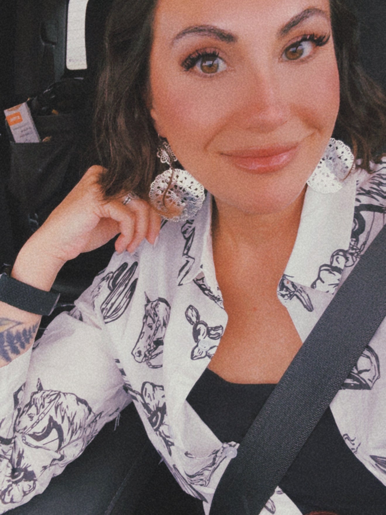 Selfie of a woman in a car wearing large silver filigree earrings, a white Western-print shirt with black horses and steer skulls, and a dark seatbelt across her chest
