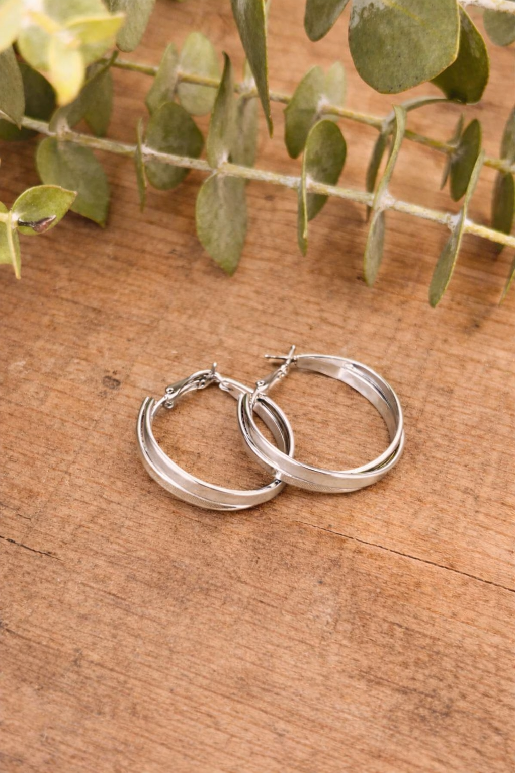 A pair of polished silver hoop earrings with a textured inner band and smooth outer band resting on a rustic wooden plank, accented by soft green eucalyptus leaves
