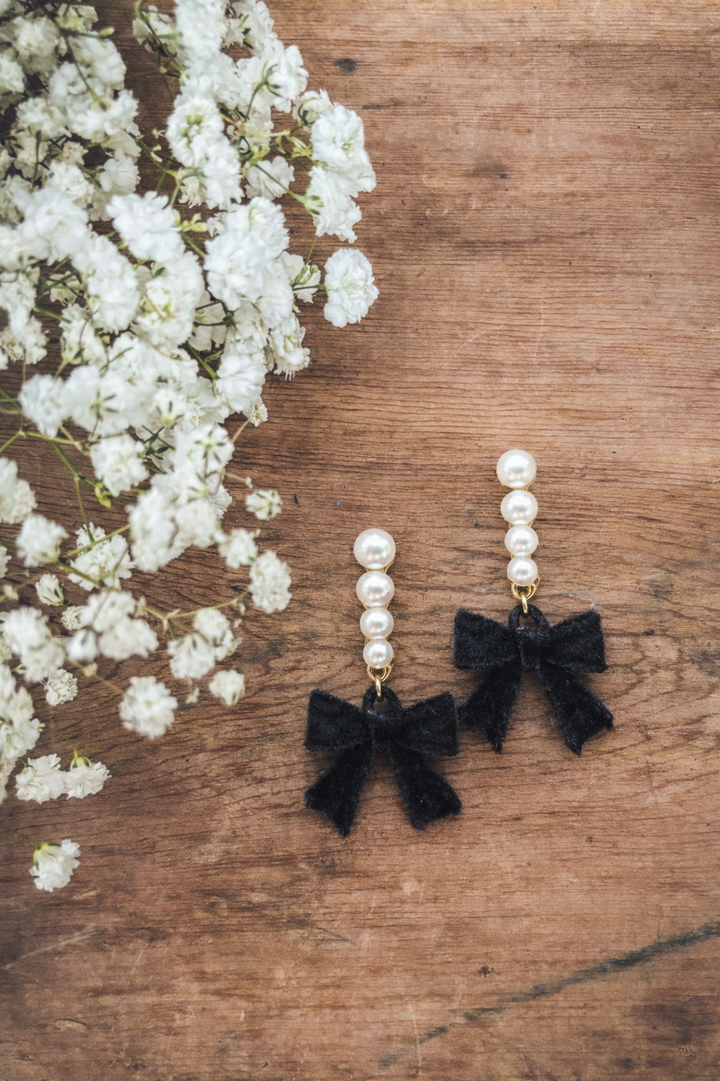 Photography of elegant dangle earrings featuring a stack of lustrous white pearls and dainty black velvet bows, displayed on a rustic wooden surface alongside delicate white baby's breath flowers
