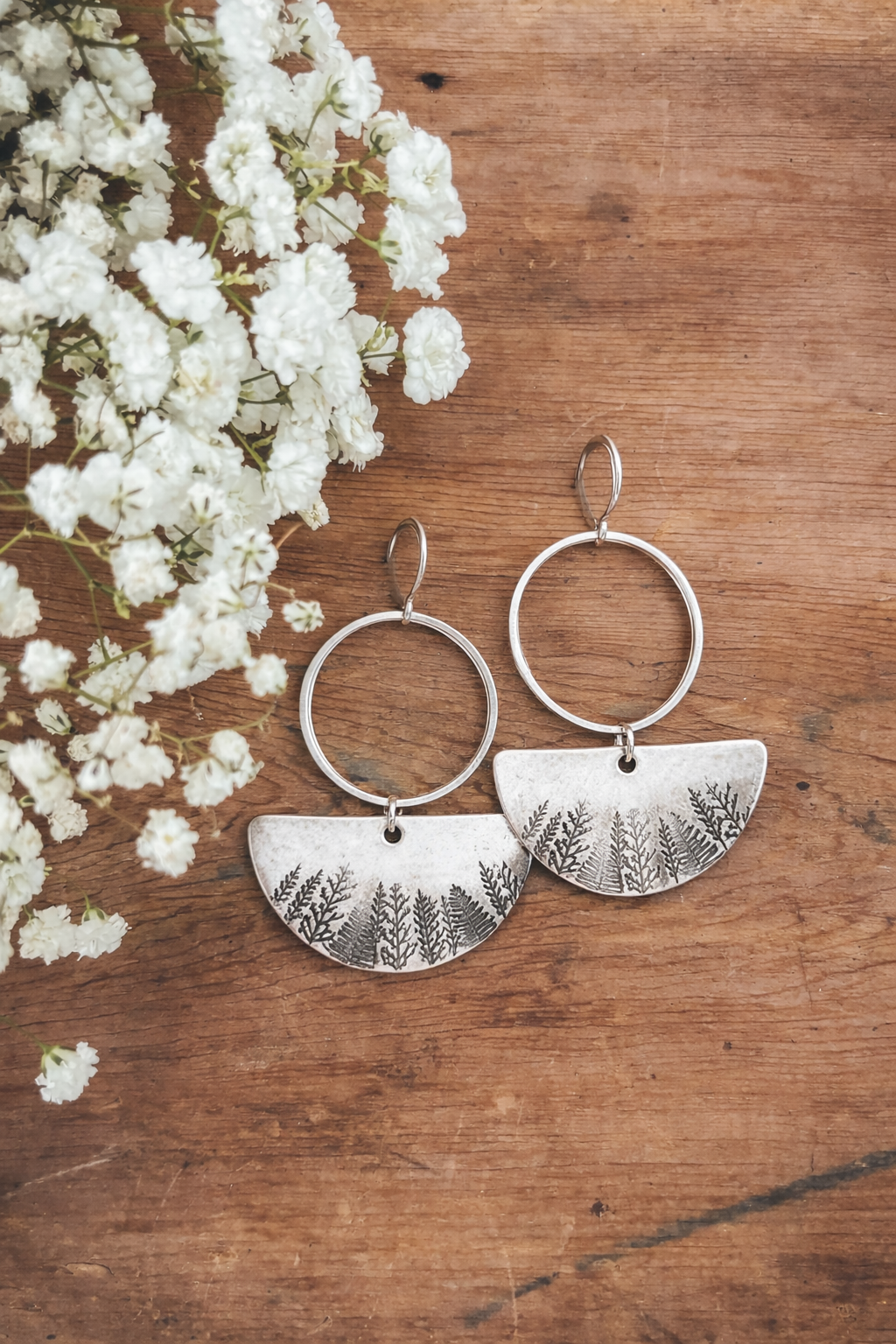 Handcrafted silver hoop earrings featuring semi-circular pendants with an etched forest pattern displayed on a rustic wooden surface alongside delicate white baby's breath flowers