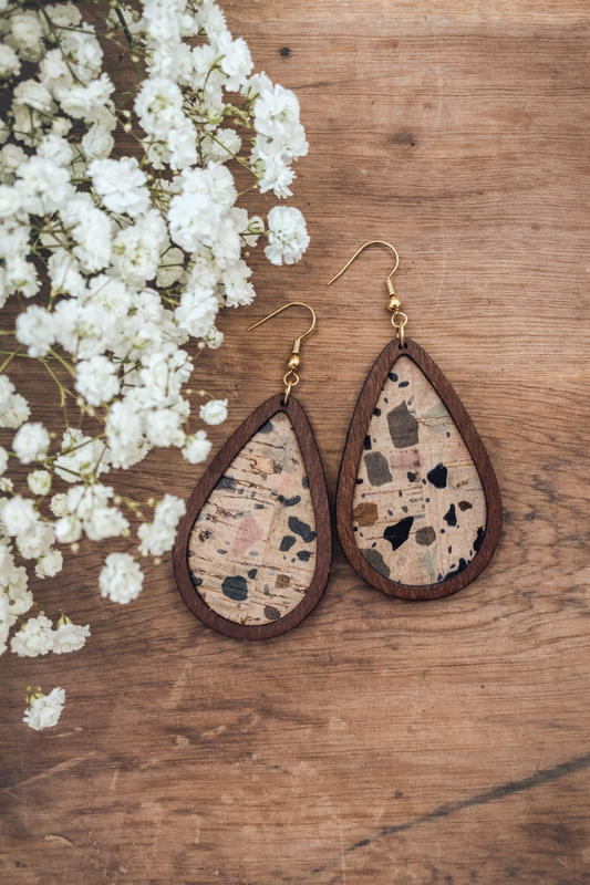 Product photography of teardrop-shaped wooden earrings with speckled cork centers featuring black, pink, and brown flecks, displayed on a rustic wooden surface next to a cluster of white baby's breath flowers