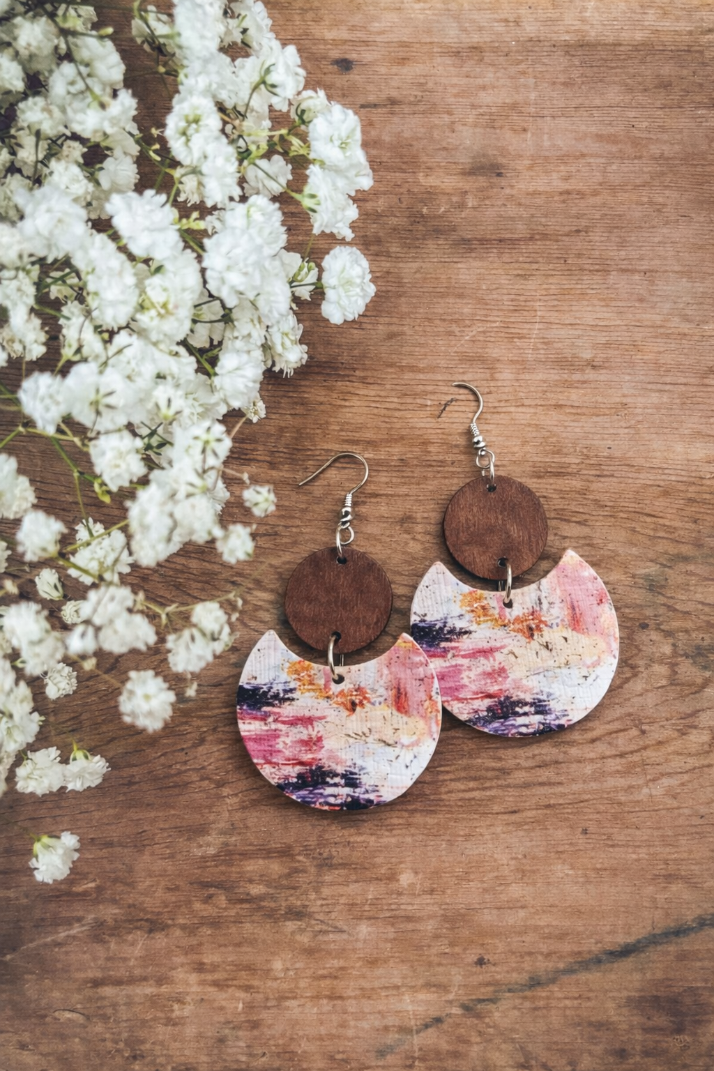 A pair of artisanal drop earrings on a rustic wooden background next to white baby's breath flowers featuring dark brown circular tops and crescent-shaped bottom pieces with abstract pink purple and orange paint strokes