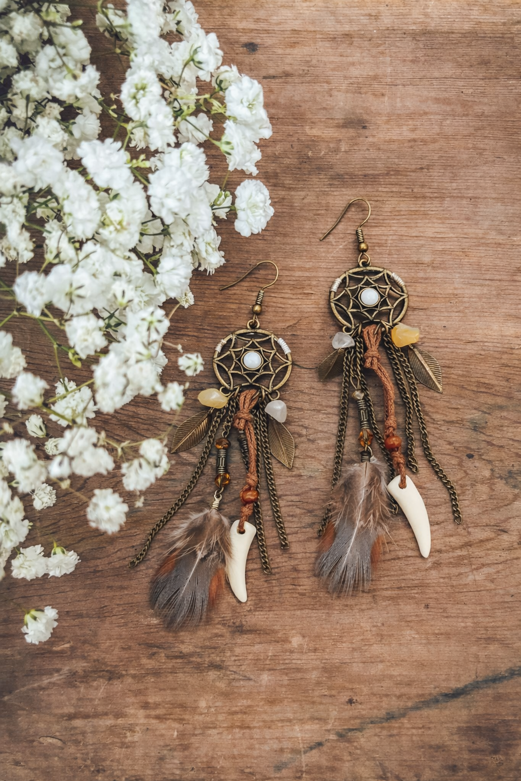 Photography of handcrafted bohemian dreamcatcher earrings featuring a central white cabochon, brass chains with leaf charms, rich brown feathers, and ivory tusk pendants, displayed on a rustic wooden surface beside delicate white baby's breath flowers