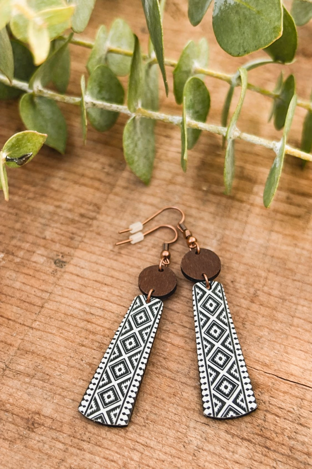 A pair of dangle earrings featuring copper hooks, dark wood circular studs, and black and white geometric patterned trapezoidal drops set against a rustic wooden background with a green eucalyptus branch