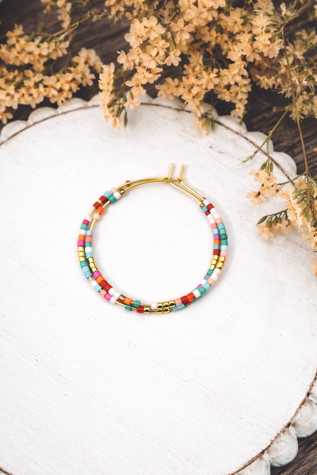 Gold-toned hoop earrings with tiny multi-colored square seed beads displayed on a white scalloped wooden tray, accented by dried light orange flowers