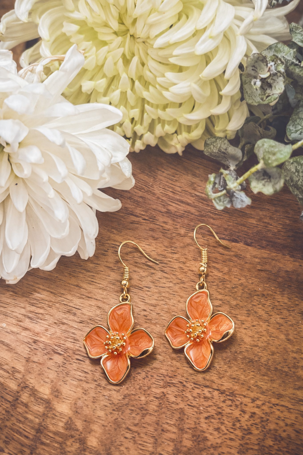 Photograph of a pair of orange enamel flower-shaped dangle earrings with gold tone details and small beaded centers on a rich brown wooden surface, framed by soft white and pale yellow chrysanthemum flowers and muted green foliage