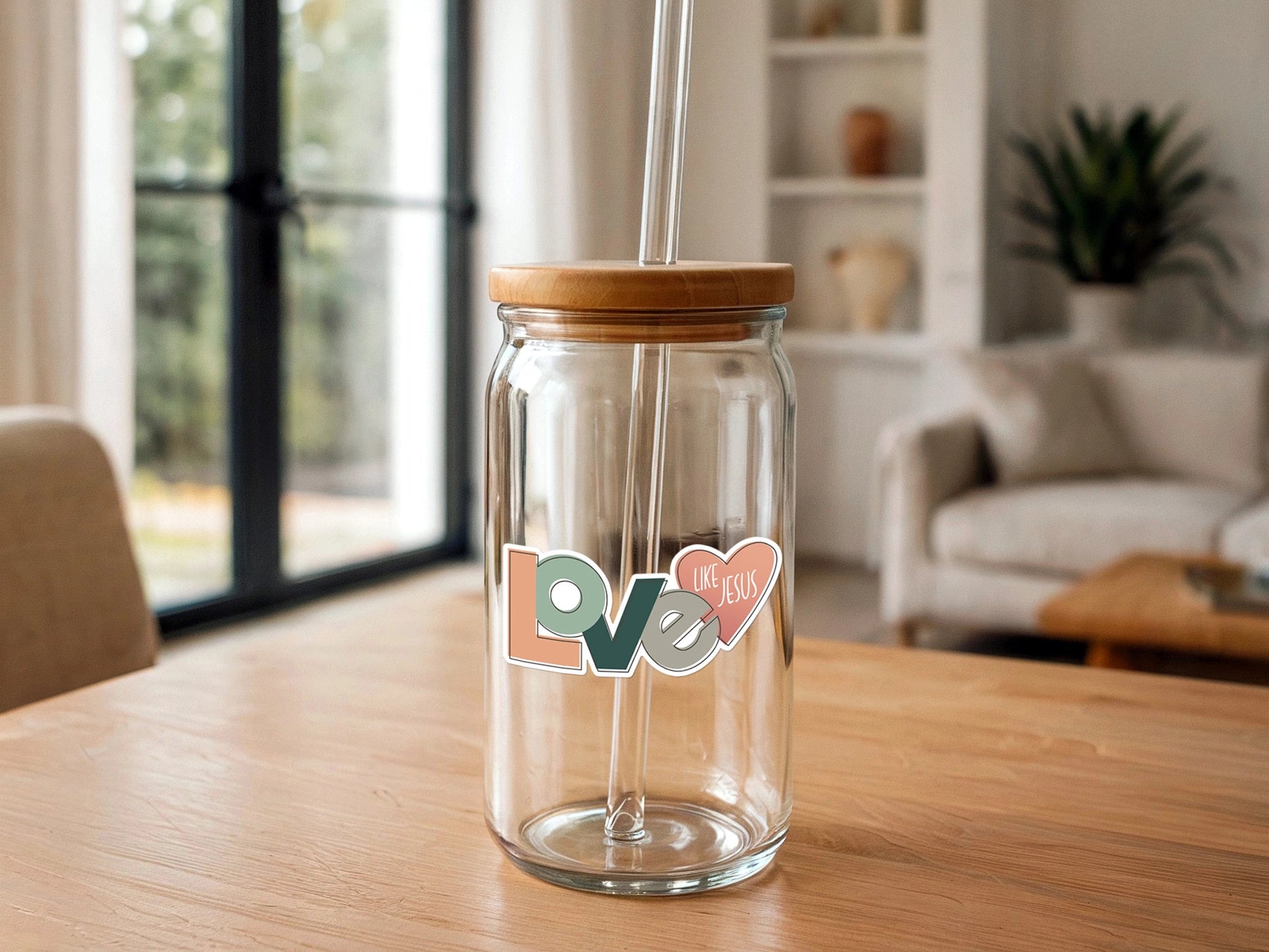Product photography of a clear glass jar with a bamboo lid and glass straw, adorned with a muted peach, green, and grey "LOVE LIKE JESUS" sticker, sitting on a light wooden table in a contemporary living room with a large window and a lush green plant