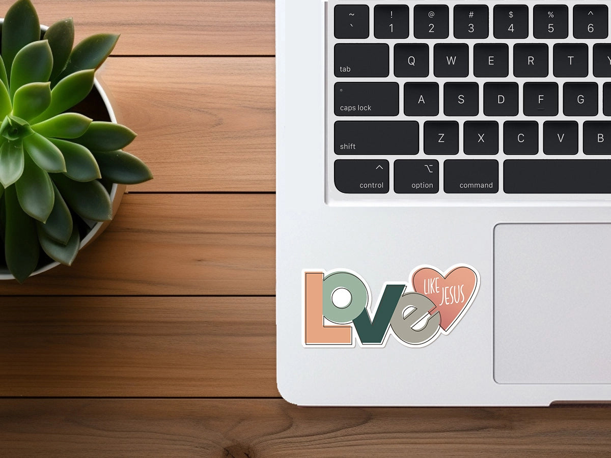Photography of a modern silver MacBook laptop on a warm wooden desk featuring a vibrant green succulent plant and two colorful stickers, one spelling LOVE in peach, teal, and gray, and a pink heart with LIKE JESUS