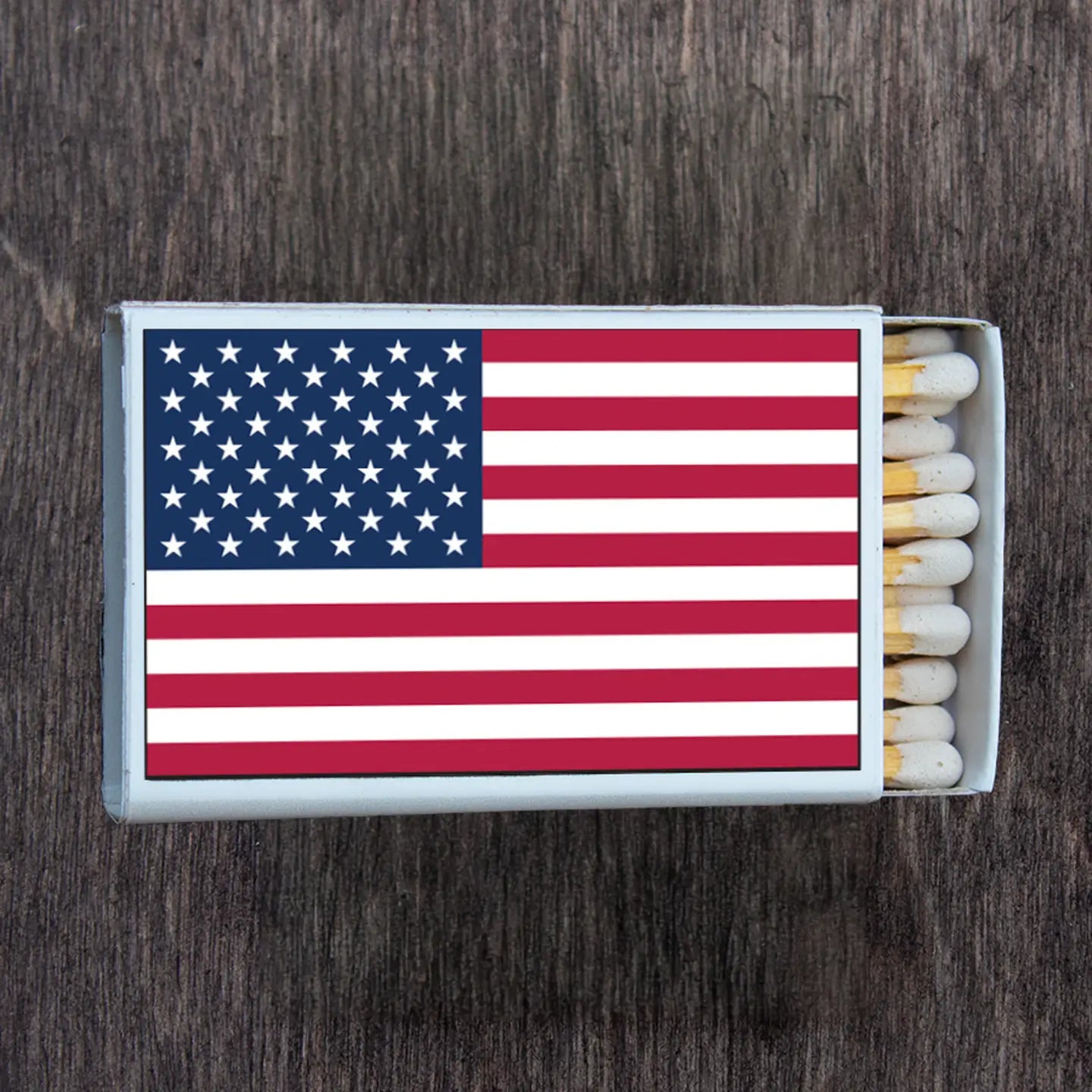A matchbox adorned with the American flag design featuring red and white stripes with a blue canton full of white stars, resting on a rustic dark wooden surface with several white-tipped wooden matches visible inside the opened box