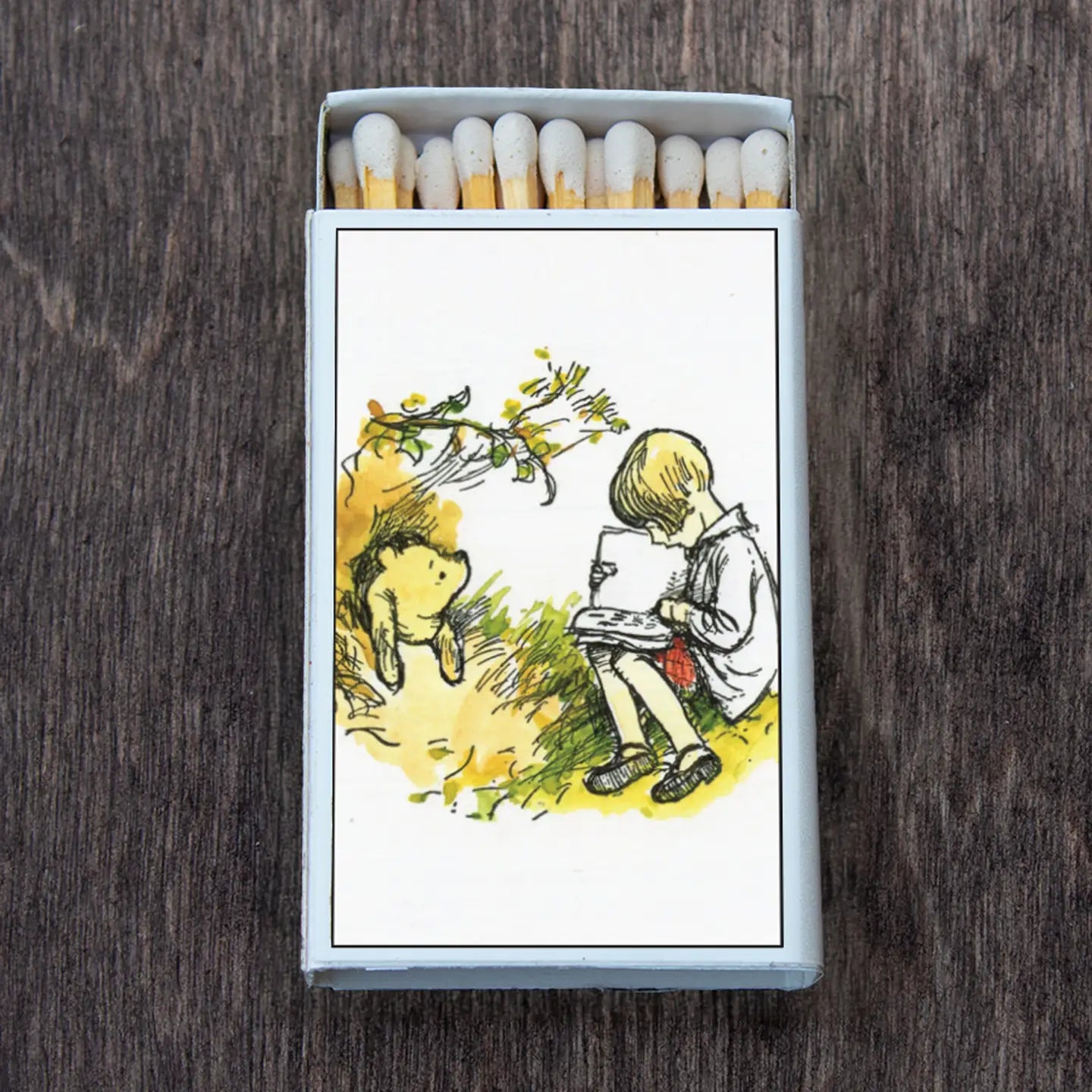 E H Shepard illustration on a matchbox showing Christopher Robin reading a book in a stylized yellow-green landscape while Winnie the Pooh peeks from behind a grassy hill
