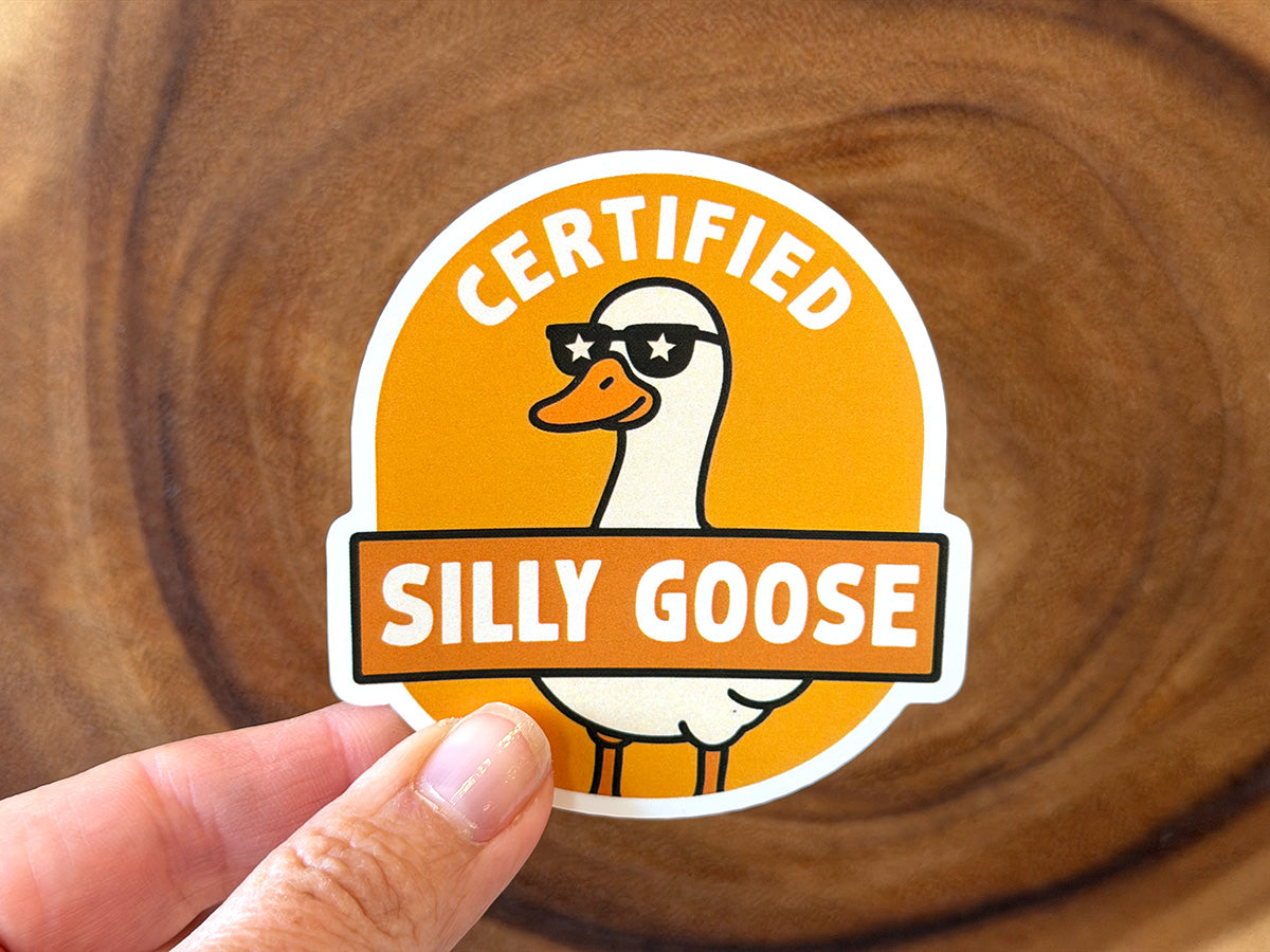 Sticker with a circular orange design depicting a white cartoon goose wearing star sunglasses and the words Certified Silly Goose held by a hand against a natural wood background