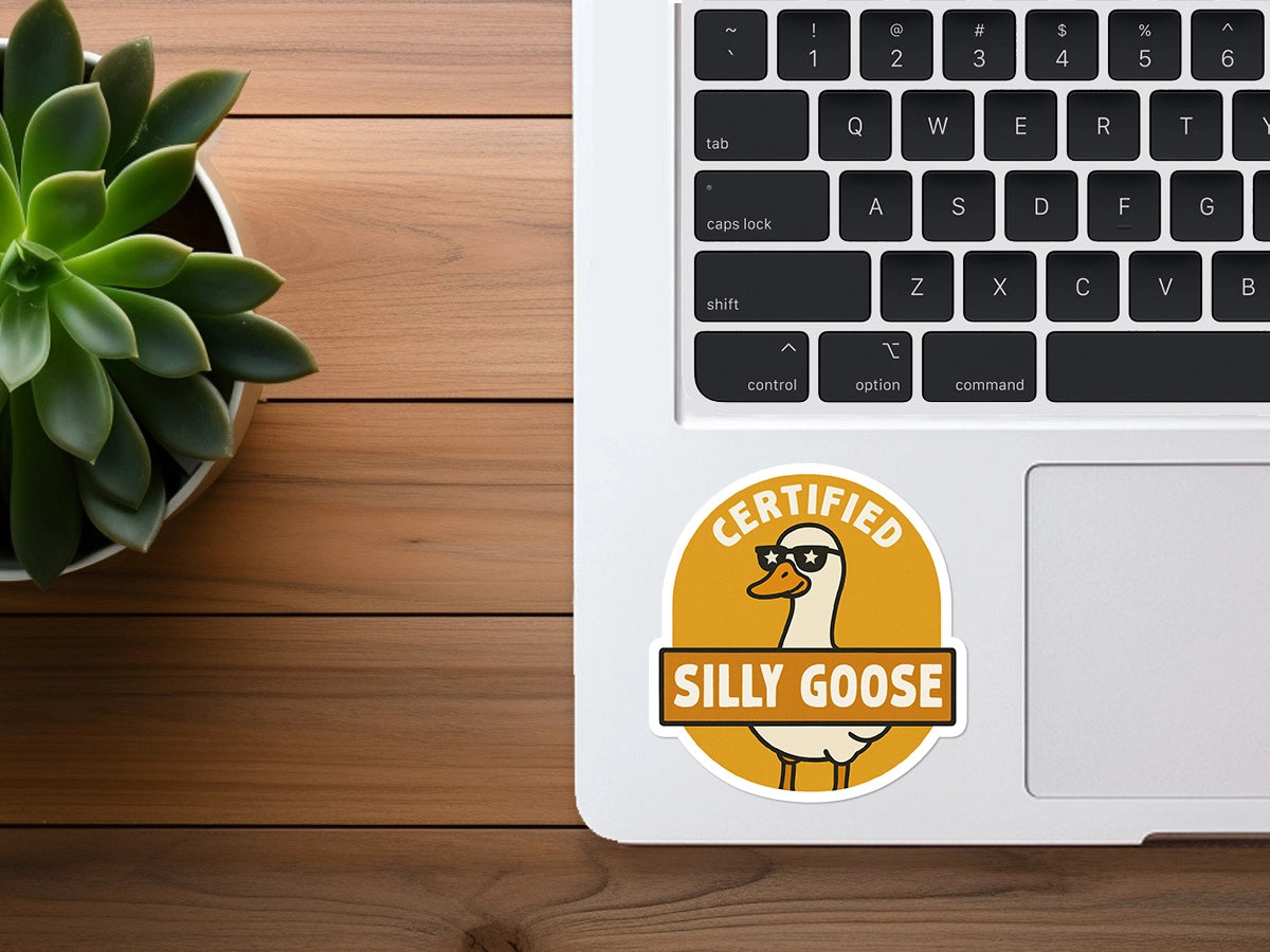 Photograph of a silver MacBook keyboard featuring a colorful round orange and yellow Certified Silly Goose sticker showing a goose in sunglasses, positioned beside a vibrant green succulent plant on a warm wooden desk