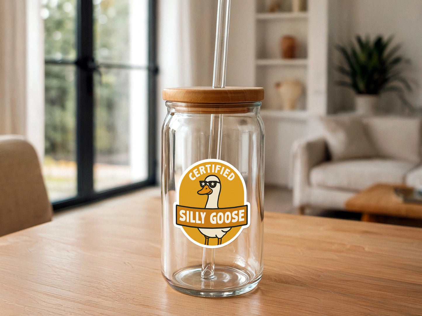 Digital photograph of a clear glass tumbler featuring a bamboo lid, glass straw, and a yellow circular "Certified Silly Goose" sticker, set on a blonde wooden table in a bright modern living room with blurred background elements like a couch and window