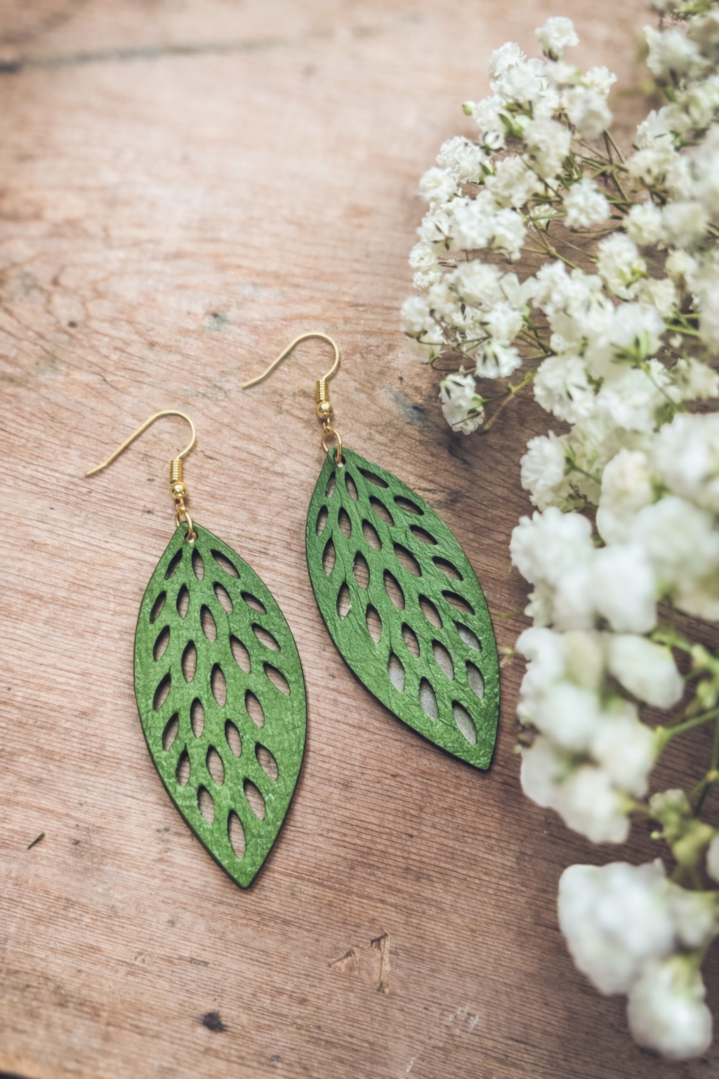 Photograph of a pair of green leaf-shaped earrings with intricate cutout patterns and gold hooks displayed on a rustic wooden surface alongside delicate white baby's breath flowers