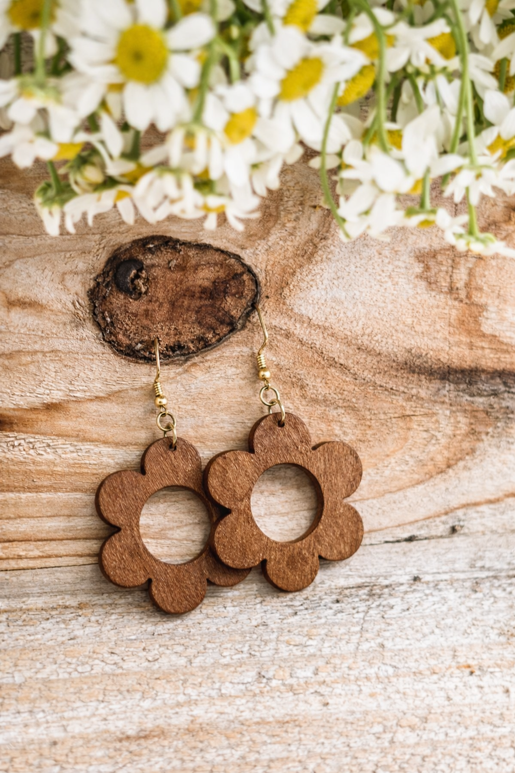 Photography showcasing a pair of dark brown flower-shaped wooden earrings with gold hooks against a rustic light wood grain background featuring a prominent dark knot, with soft focus white and yellow daisies at the top