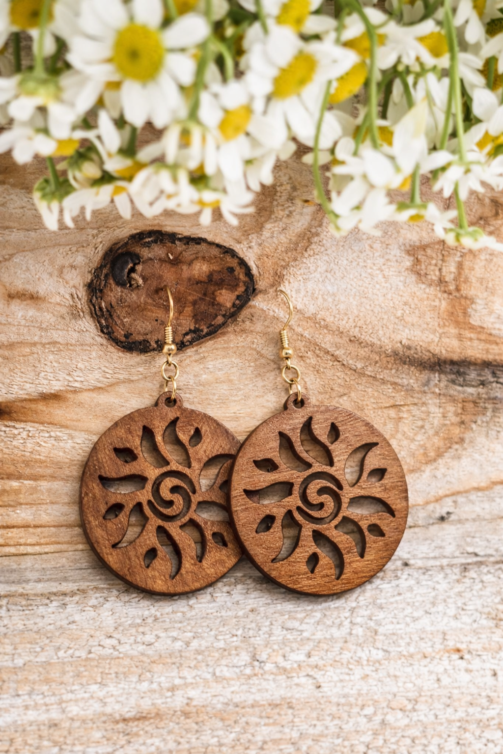 Photography of a pair of round dark brown wooden earrings with intricate laser-cut sun and spiral designs and gold hooks, displayed on a rustic light-grained wooden surface with a prominent dark knot, beneath blurred white and yellow daisy blossoms