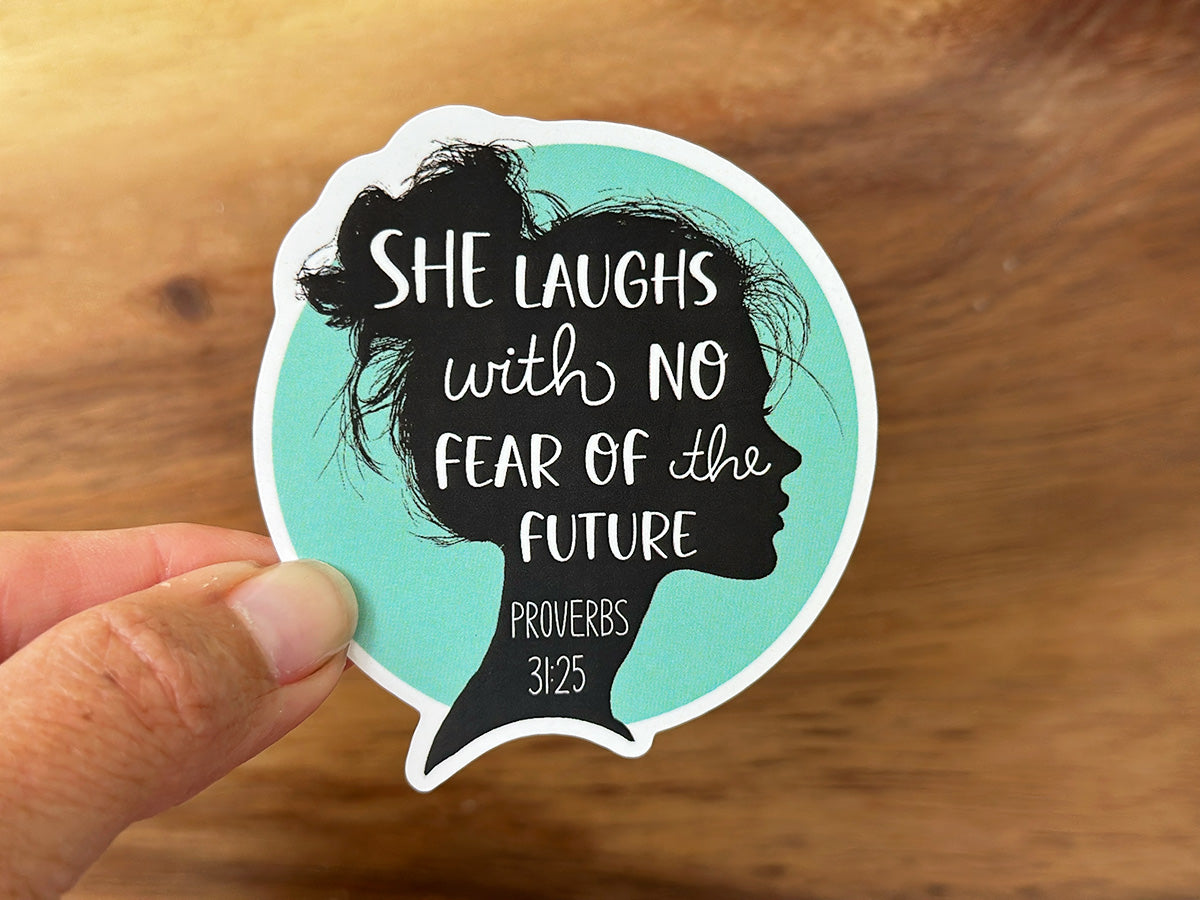 Die-cut sticker depicting a black female head silhouette with a messy bun on a teal background, featuring white script text SHE LAUGHS with NO FEAR of the FUTURE PROVERBS 31:25, held by fingers against a warm wooden table