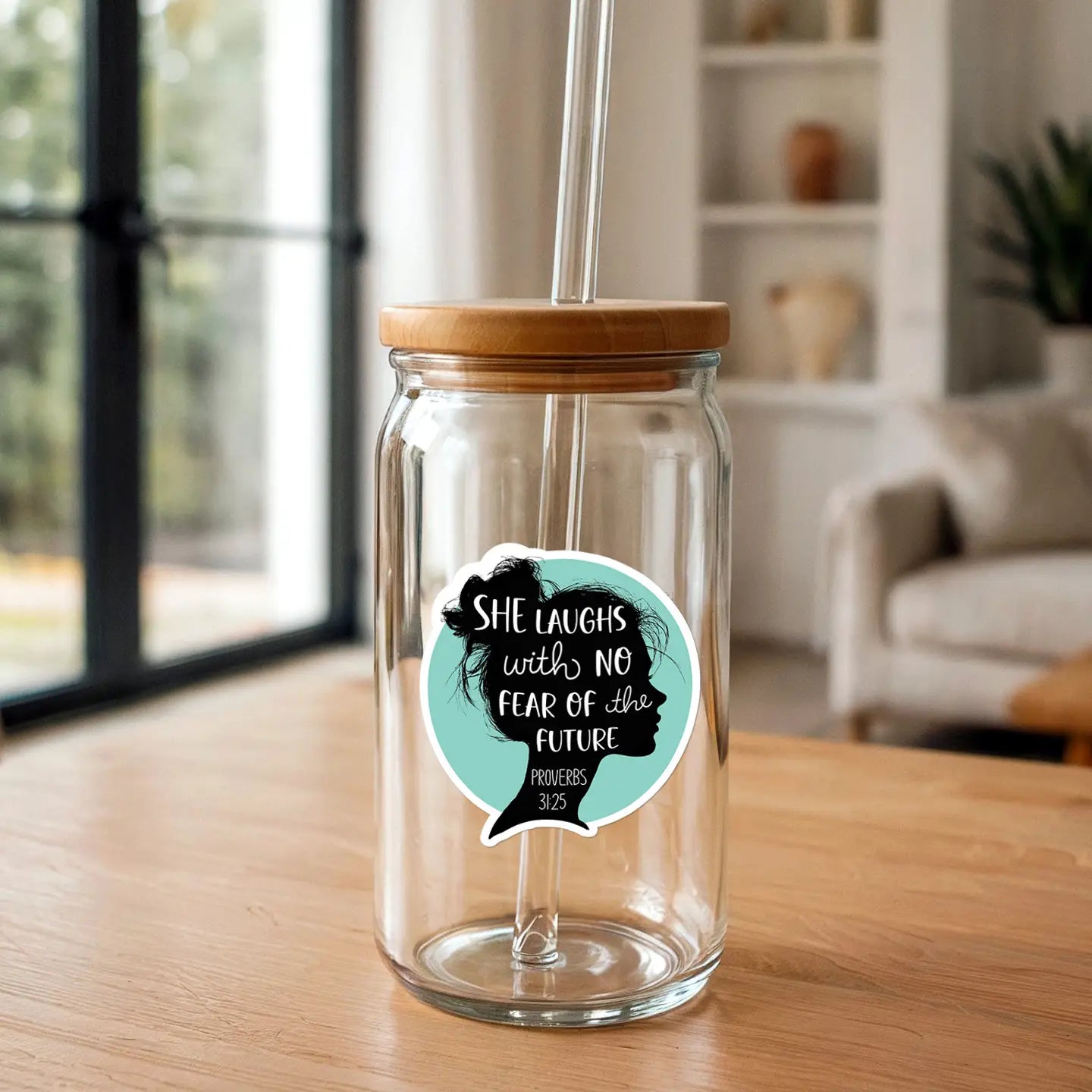 Clear glass tumbler with a bamboo lid and glass straw featuring a turquoise sticker with a woman's silhouette and the quote 'SHE LAUGHS WITH NO FEAR OF THE FUTURE PROVERBS 31:25' on a light wooden table in a modern home