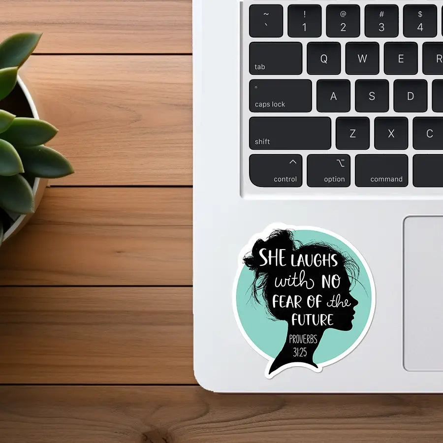 Sticker art depicting a circular teal sticker with a black silhouette of a woman in profile, text reading "SHE LAUGHS with NO FEAR of the FUTURE PROVERBS 31:25", placed on a white laptop keyboard next to a light wooden desk and a green succulent plant