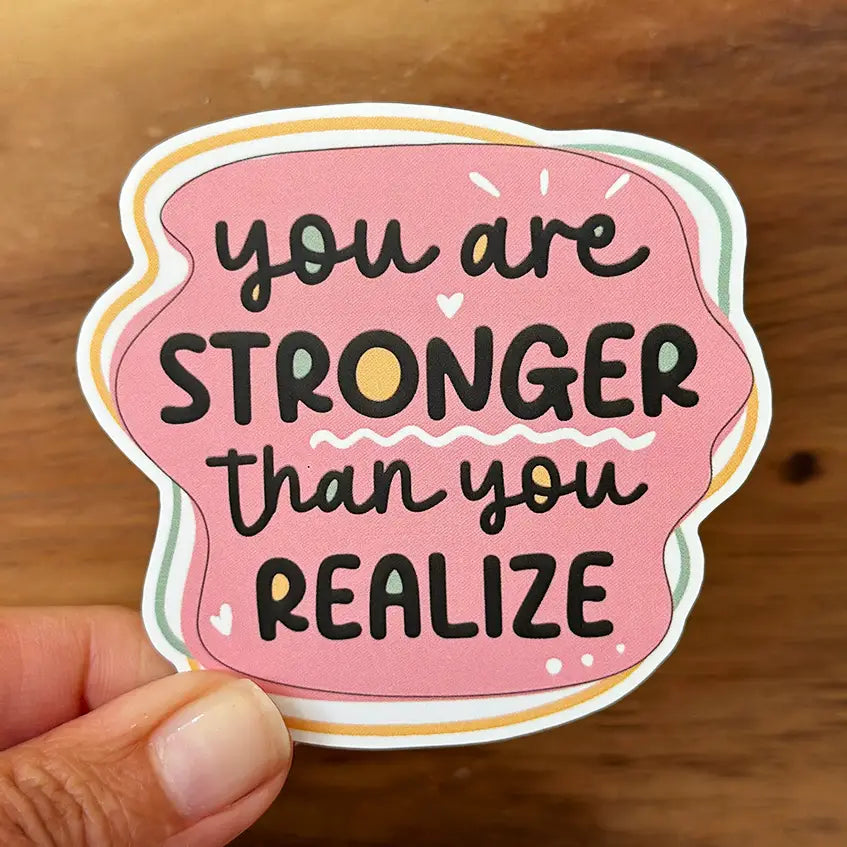 Sticker featuring the inspirational quote You are STRONGER than you REALIZE in black text with a yellow O on a pink background, outlined in pastel green yellow and white, and held by a hand against a wooden surface