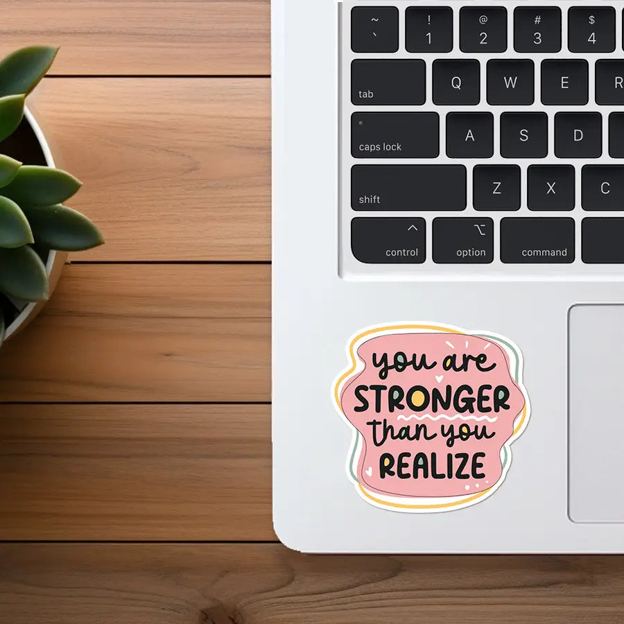 Photography of a "you are STRONGER than you REALIZE" motivational sticker with a pink, yellow, and light green border on a white MacBook laptop next to a green succulent plant in a white pot on a wooden desk