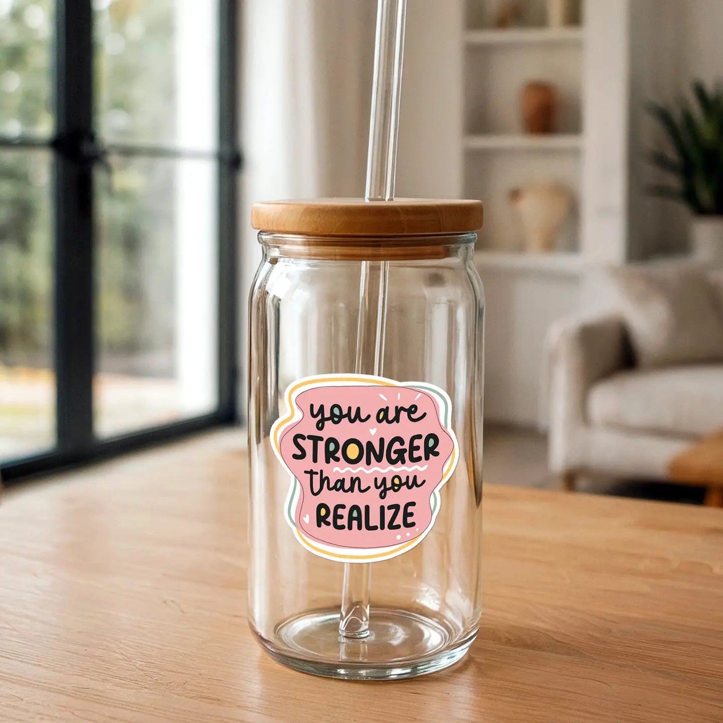 A clear glass drinking jar with a bamboo lid and glass straw, featuring a pink sticker reading "you are stronger than you realize", sits on a light wooden table in a blurred modern indoor background with a window and white bookshelf
