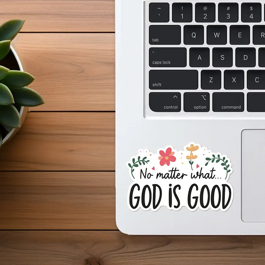 Photograph of a white MacBook laptop featuring a colorful sticker that reads No matter what... GOD IS GOOD with pink and yellow flowers and green leaves, set on a wooden desk beside a green succulent plant