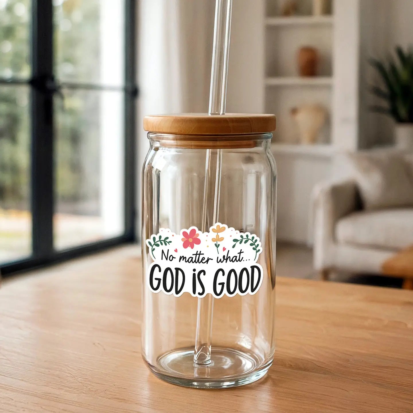 Photography of a clear glass tumbler with a bamboo lid and glass straw featuring a sticker with pink and yellow floral accents and the text No matter what GOD IS GOOD on a light wooden table indoors