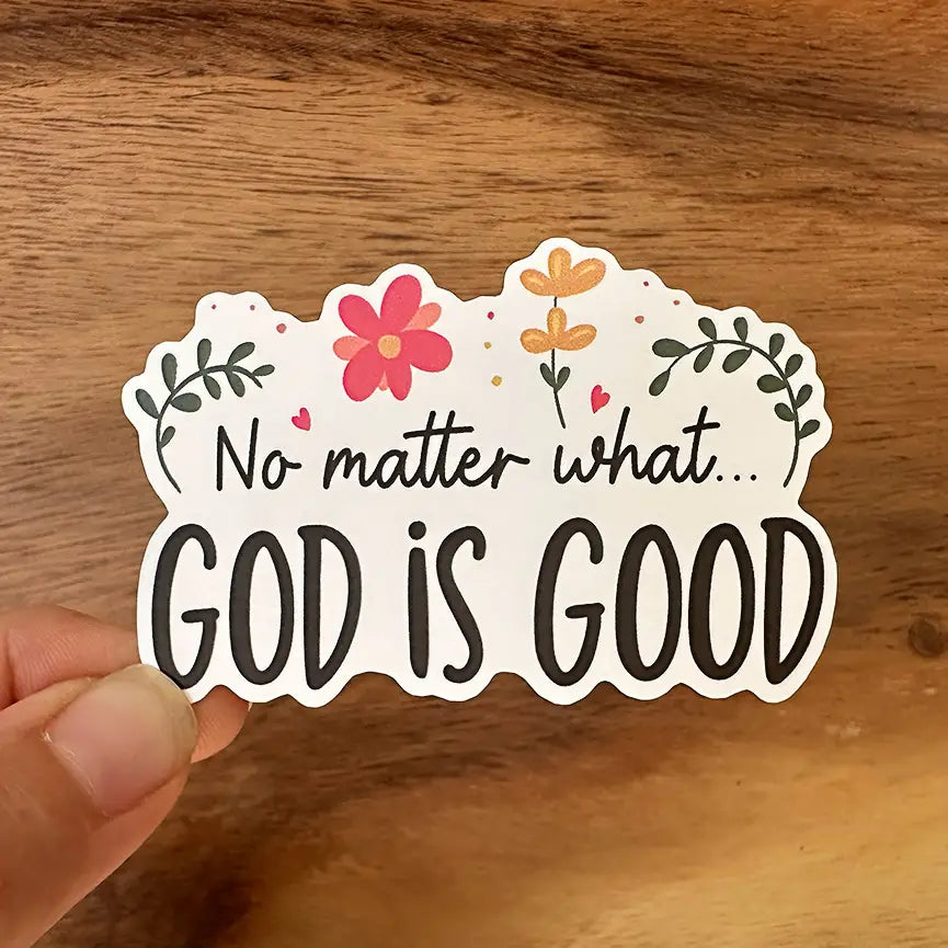 Die-cut sticker featuring the phrase No matter what GOD IS GOOD in black script, adorned with pink and orange flowers, green leaves, small red hearts, and gold glitter accents, held by a hand against a wooden background